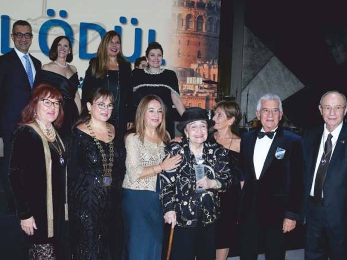 Nurdan Üstman, con sombrero negro, sostiene en el escenario el premio Lifetime Achievement de Skål International, rodeada de socios en traje de noche